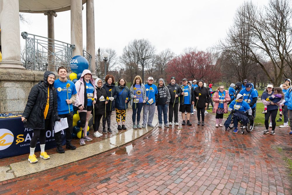 Group of runners gathered under a gazebo on wet brick pathway during rainy weather wearing blue athletic gear.