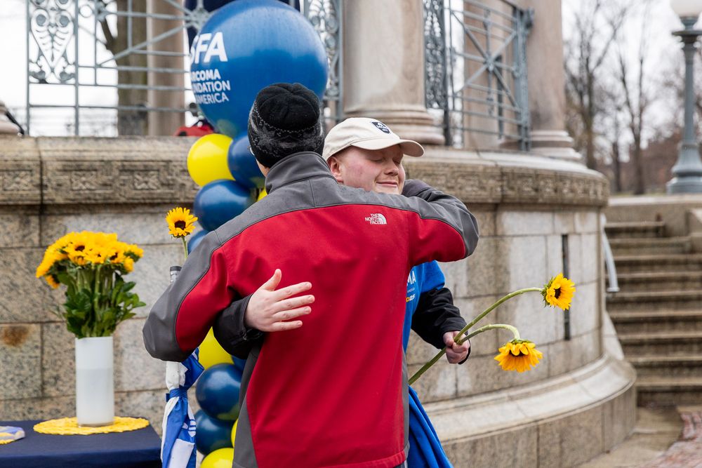 A person in a red sweatshirt embraces a blue balloon mascot in front of stone steps with yellow flowers nearby.