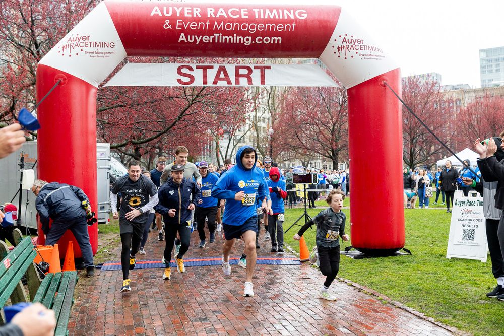 Runners take off from a red inflatable START arch at an outdoor race event on a wet spring day with cherry blossoms visible.