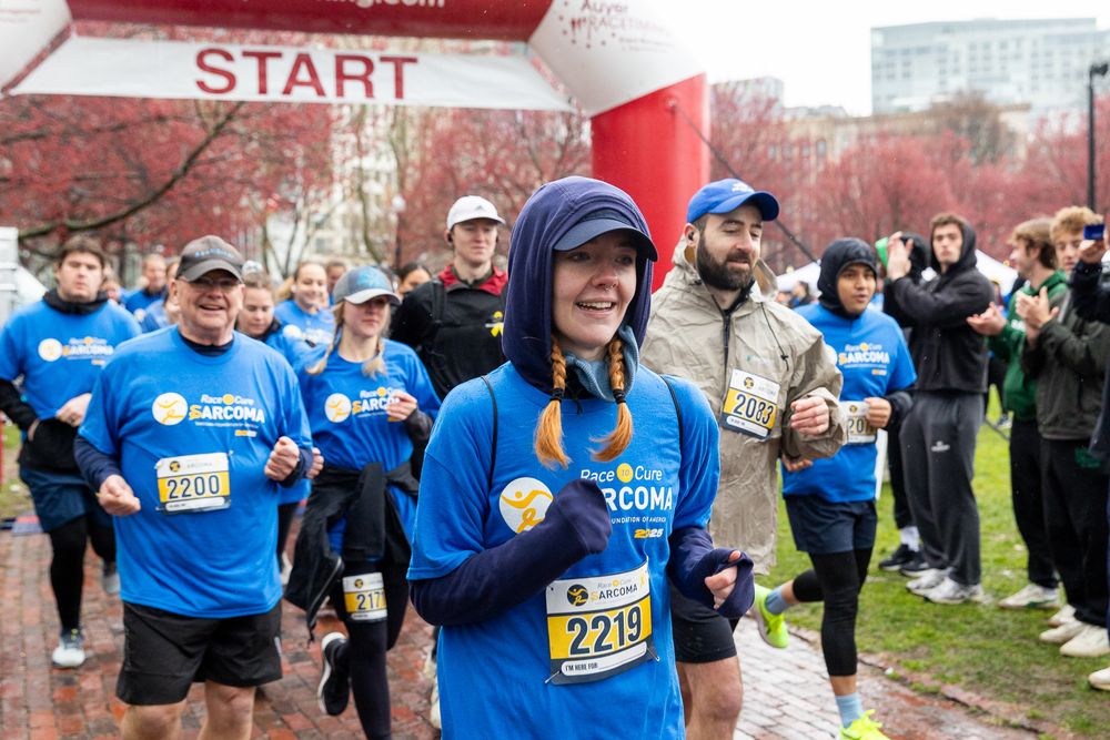 Marathon runners in blue jerseys compete in a race as spectators watch from the sidelines on a cloudy day.