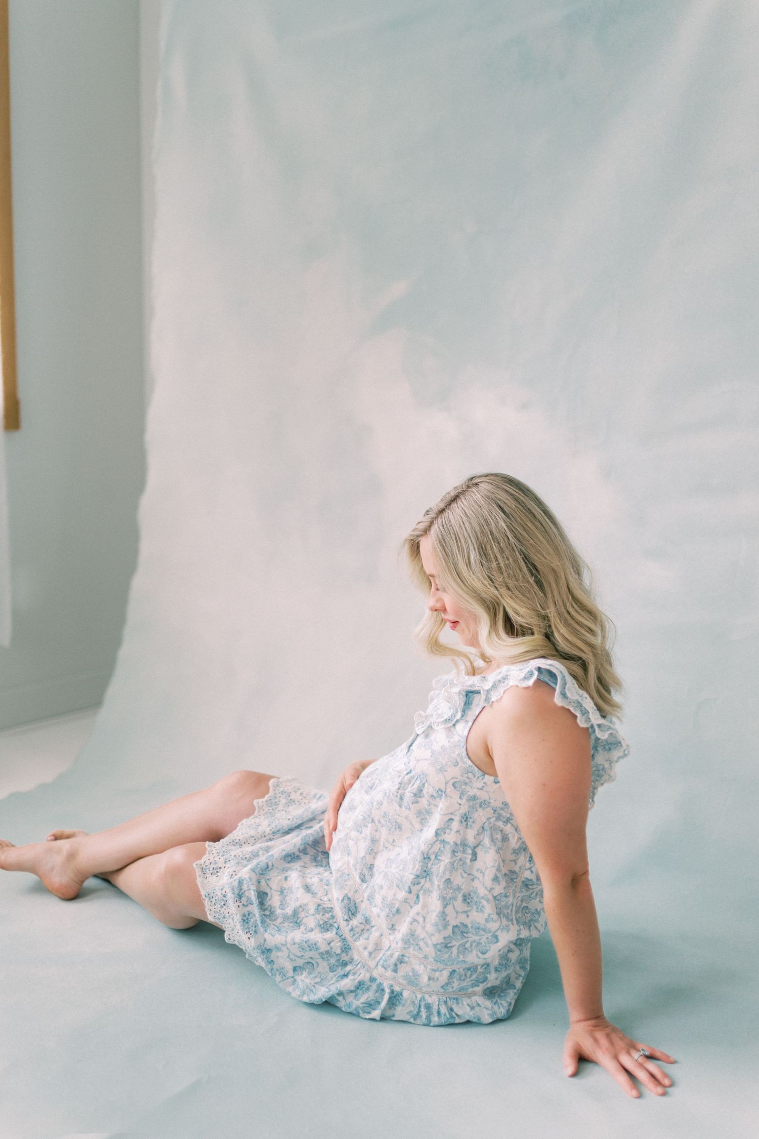 A person in a light floral dress sits on a pale blue floor against a white wall in soft natural lighting.
