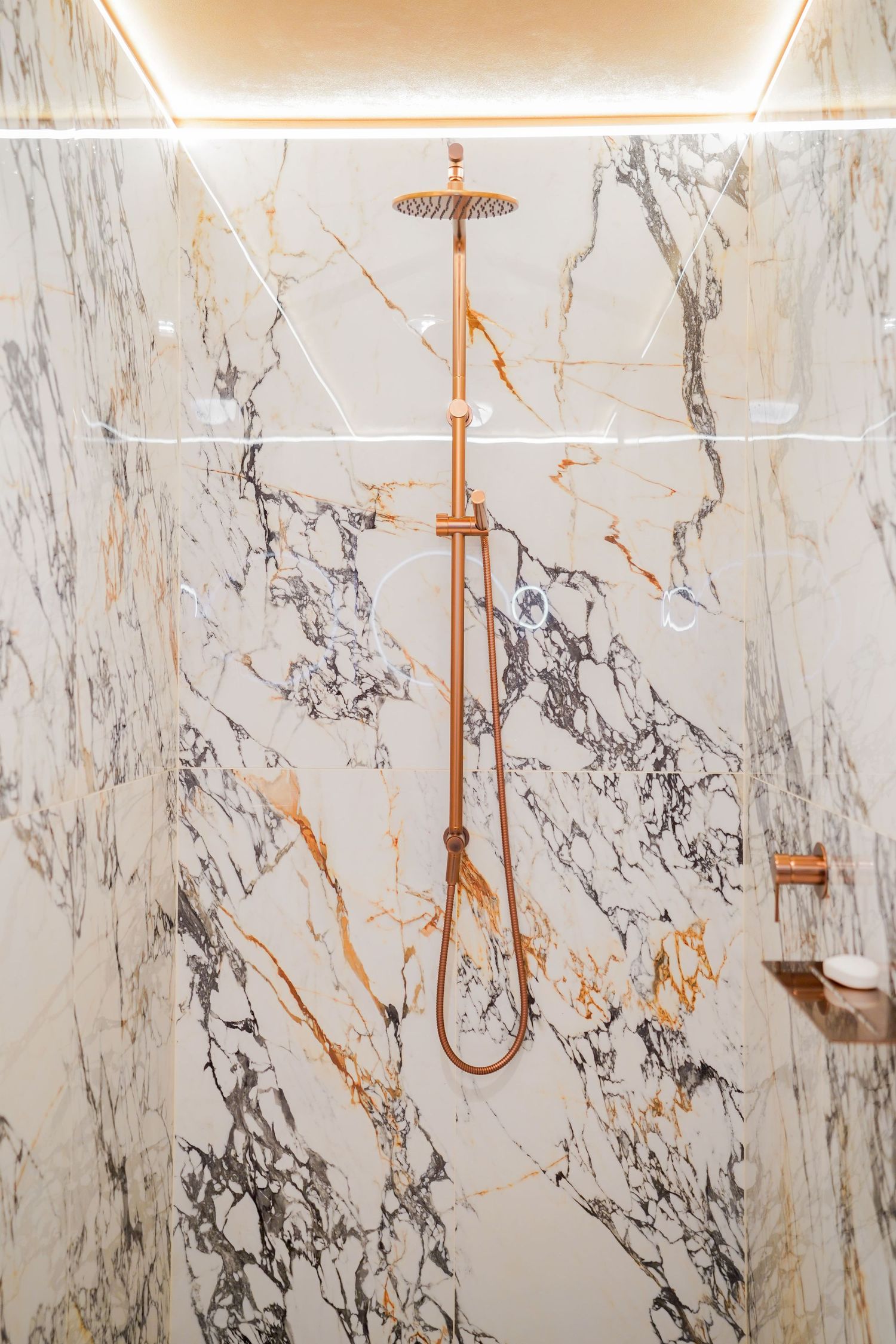 Luxurious marble bathroom shower with copper fixtures and modern lighting installed in white and grey veined stone walls.