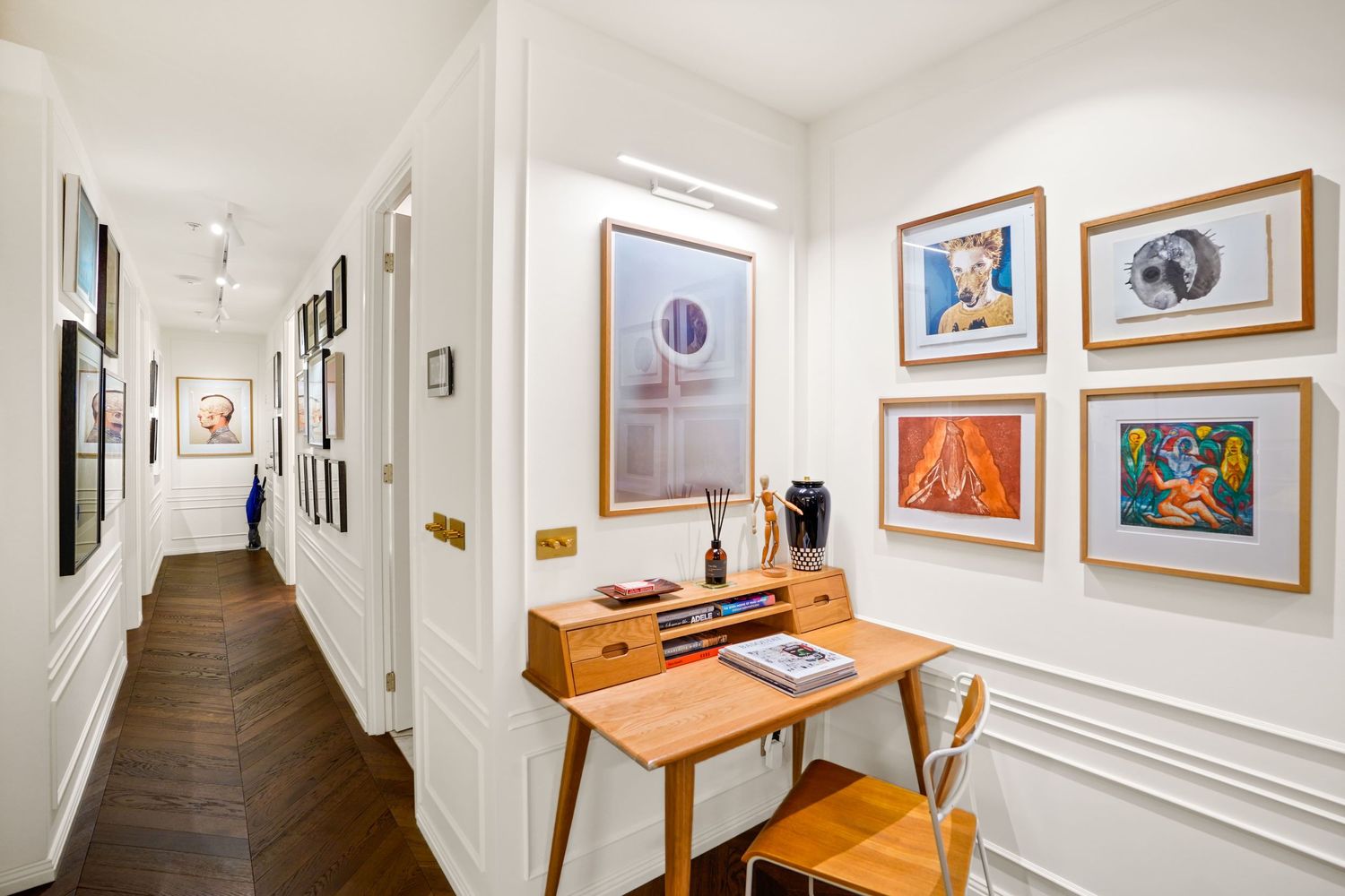 Long white hallway with wooden flooring and art gallery walls featuring colorful framed artwork above an antique desk and chair.
