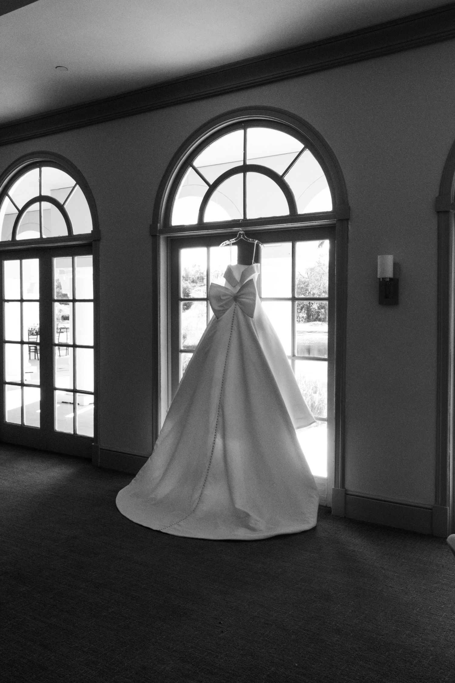 A wedding dress hangs elegantly in front of arched windows in dramatic black and white.