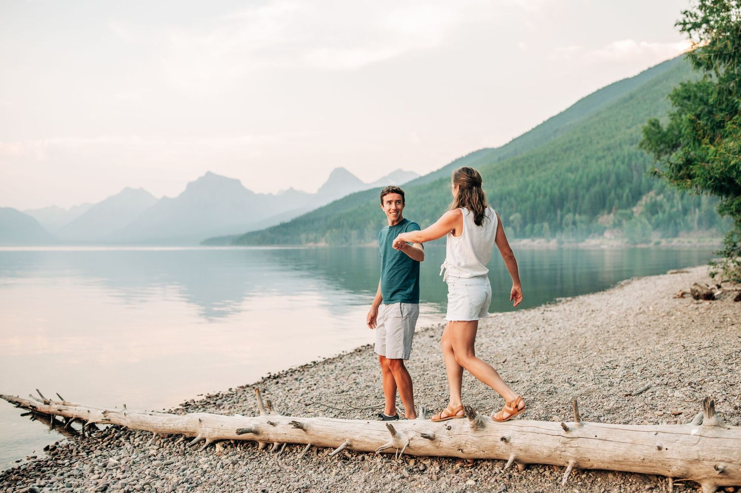 Couple walking together on fallen log by a smokey lake with mountains in background at sunset.