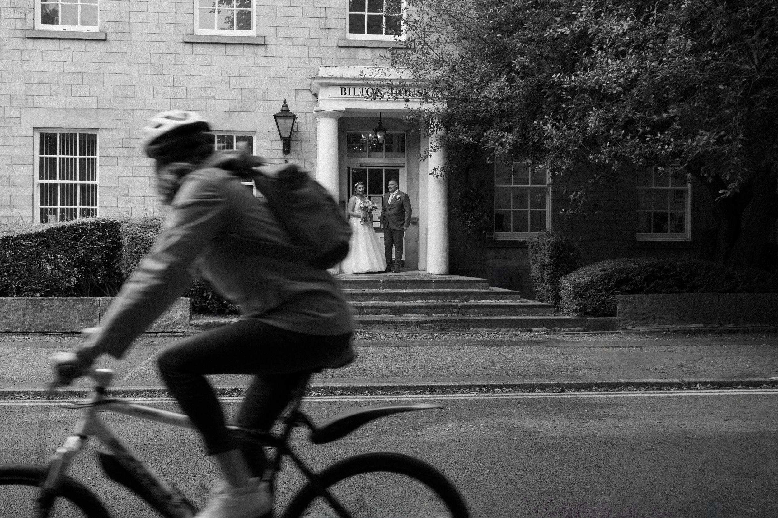 Bride & groom at registry office in Harrogate after getting married as cyclist passes in front of photographer. black white documentary