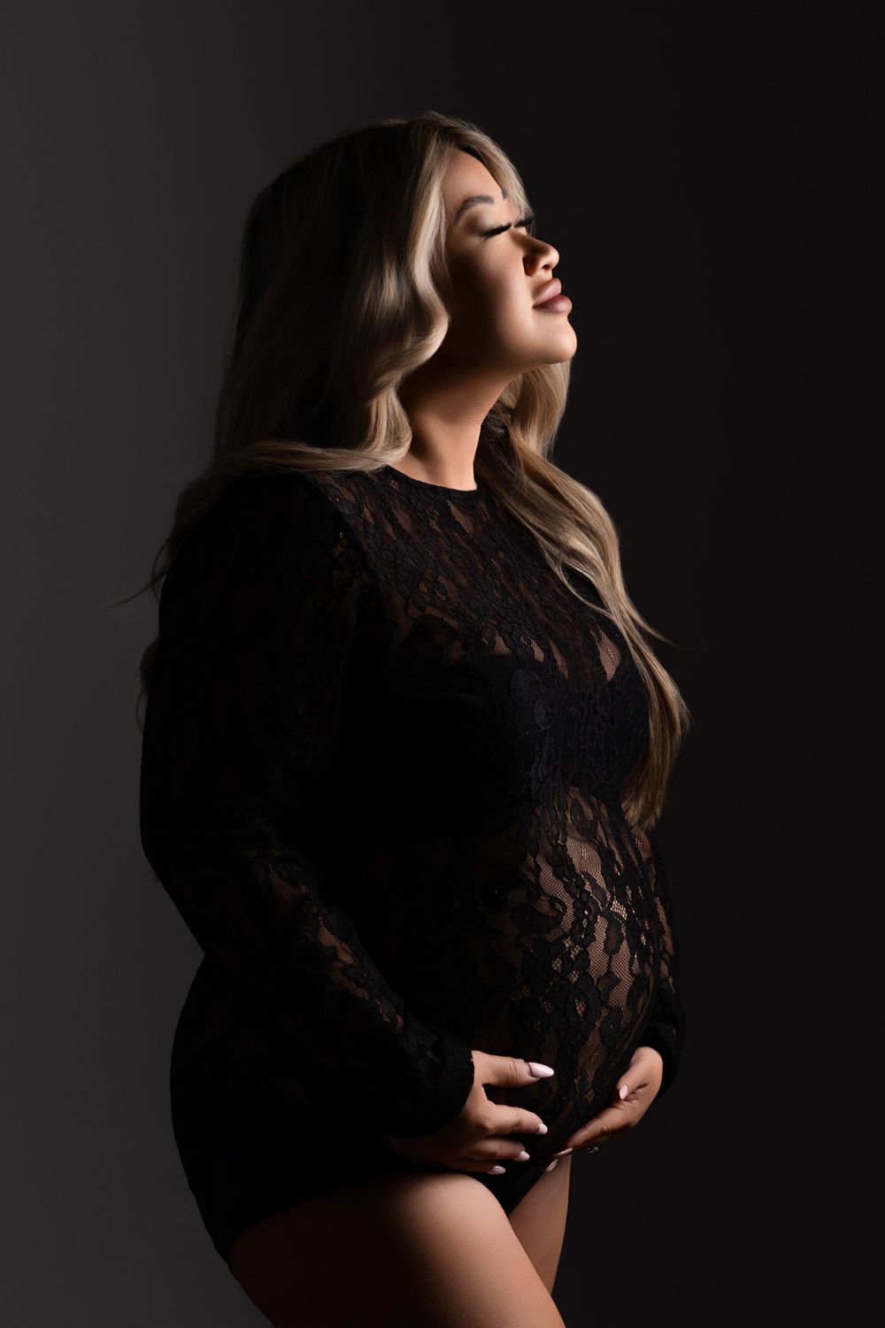 Artistic portrait of someone in black lace bodysuit posing against dark background with dramatic side lighting.