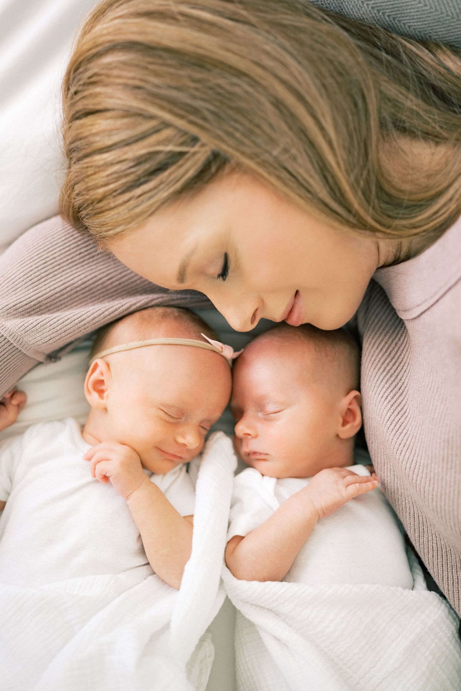 A tender moment between twins in white clothing and someone lying close to them in a beige sweater.