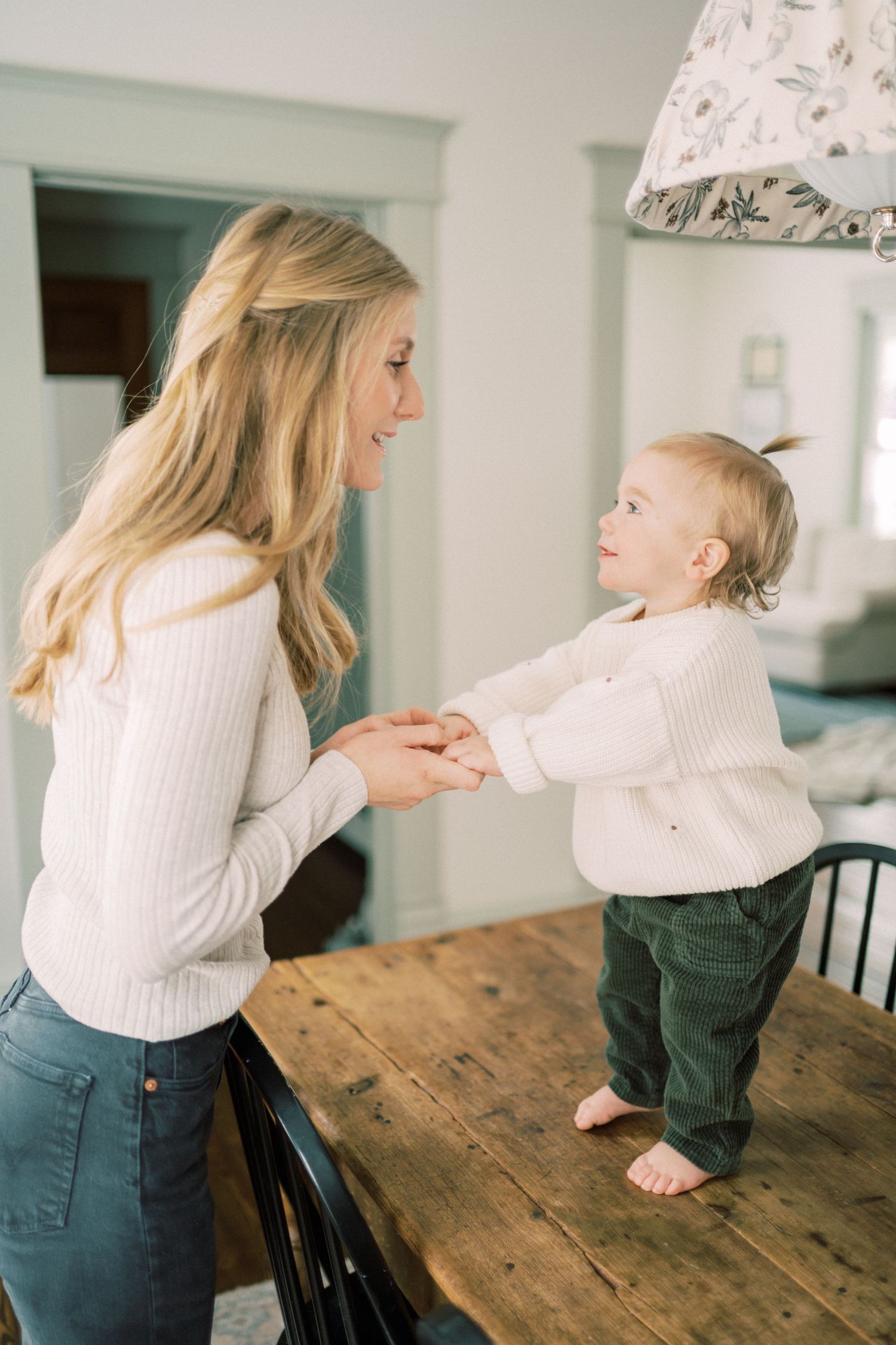 Parent and child hold hands while standing on rustic wooden table in bright home interior.