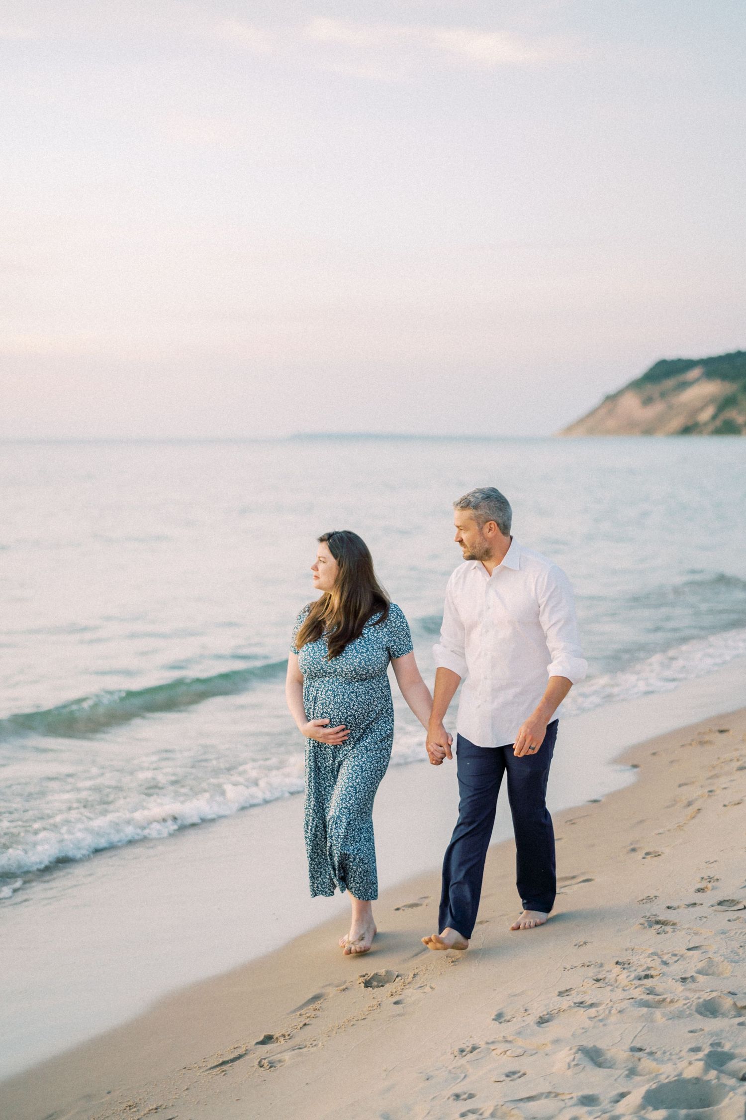 Expectant couple walking hand in hand along peaceful beach at sunset.