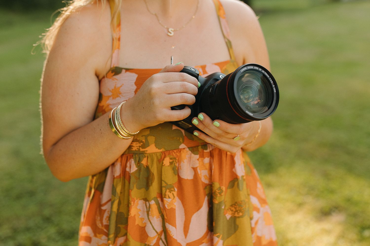 Someone wearing an orange floral dress holds a professional DSLR camera with a large lens outdoors in sunlit grass.