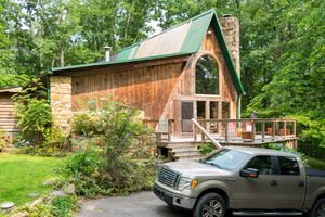Panoramic view of a rustic wooden cabin surrounded by green trees and parked vehicles.