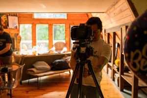 Videographer operates a professional camera on tripod in sunlit room with wooden interior.