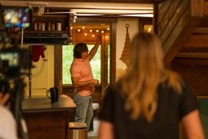 Film crew member in pink shirt working on set with string lights visible in background.
