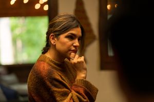 A person in a brown sweater sits thoughtfully in warm indoor lighting with a contemplative expression.