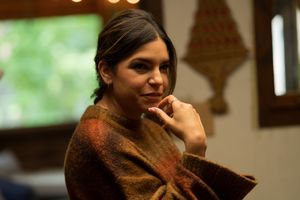 A person in a brown sweater smiles warmly in an interior setting with wooden decor.