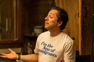 Person in white t-shirt with text making expressive hand gesture indoors.