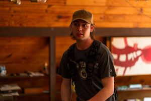 A person wearing a baseball cap stands in a wooden interior with dramatic lighting.