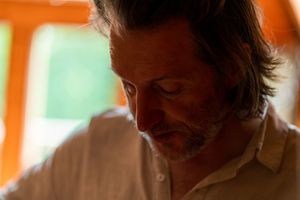 A person with facial hair looks down thoughtfully in warm orange lighting.