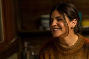 A person in a brown sweater smiles brightly in warm indoor lighting.