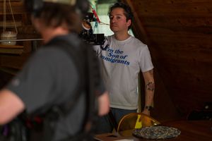 Camera crew films someone wearing a white t-shirt with text in what appears to be a dimly lit interior setting.