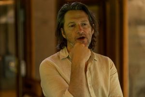 A person in a light colored shirt looks contemplative while resting their hand near their chin in an indoor setting.