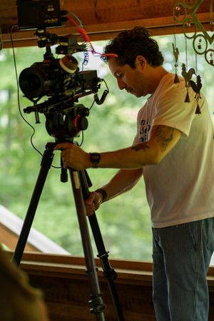 Camera operator adjusts professional video equipment on tripod while working in a natural-lit indoor setting.