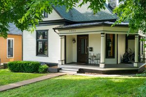 Charming white Victorian house with green trim features wraparound porch, manicured lawn and mature trees.