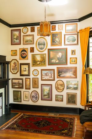 A vintage gallery wall filled with ornate frames displaying artwork and paintings above a red oriental rug on hardwood floors.