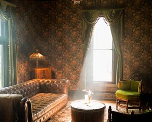 A vintage Victorian living room with floral wallpaper, tufted leather sofa, green chair and sunlit window with elegant drapes.