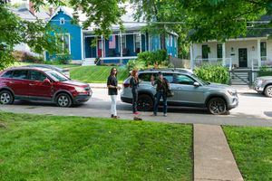 People gathered around cars on a residential street with a bright blue house in the background.