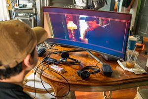 Gamer watches screen while gaming peripherals and equipment rest on wooden desk with curved monitor displaying gameplay.