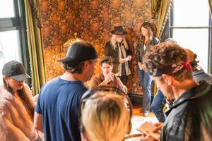Film crew and cast members gather around in a vintage-styled room with ornate wallpaper during a production meeting.