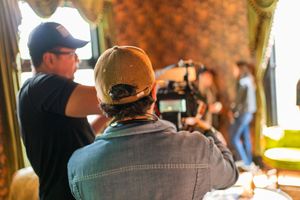 Camera operators wearing baseball caps film a scene in a room with floral wallpaper.