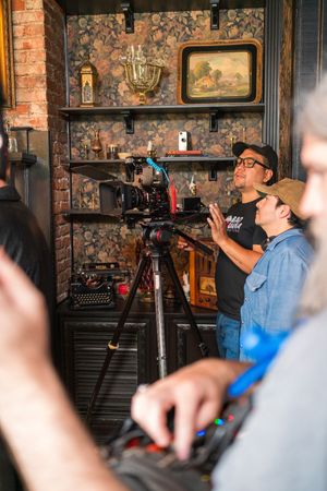Camera operators film a scene using professional equipment against a vintage wallpapered wall with decorative shelving.