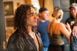 Person with long dark curly hair in a black shirt looking to the side in an indoor setting.