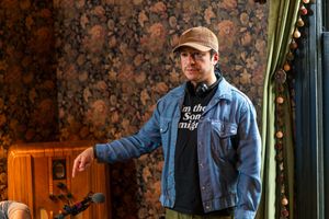 Filmmaker in denim jacket and baseball cap stands against patterned wall with camera equipment.