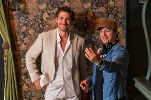 Two men in a room with ornate floral wallpaper, one wearing a light suit and the other in casual denim.