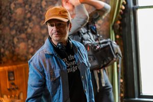 Film crew member in orange cap and denim jacket poses on set with camera gear visible.