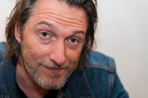 Close up portrait of a person with shoulder-length hair wearing a denim jacket against a white background.