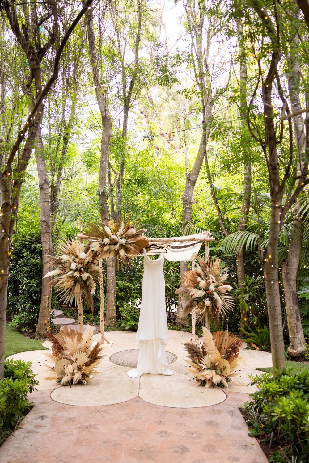 A boho wedding arch decorated with dried palm leaves and white fabric stands in a lush garden setting with tall trees.