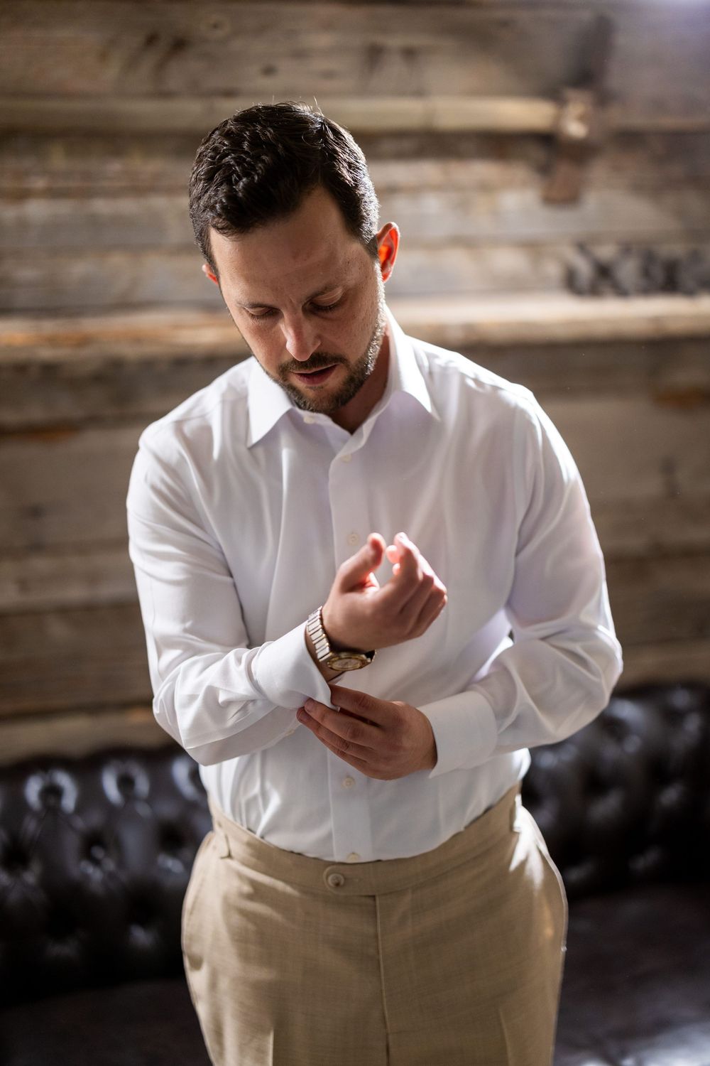 Person in white dress shirt and khaki pants adjusting sleeve cuff in rustic indoor setting.