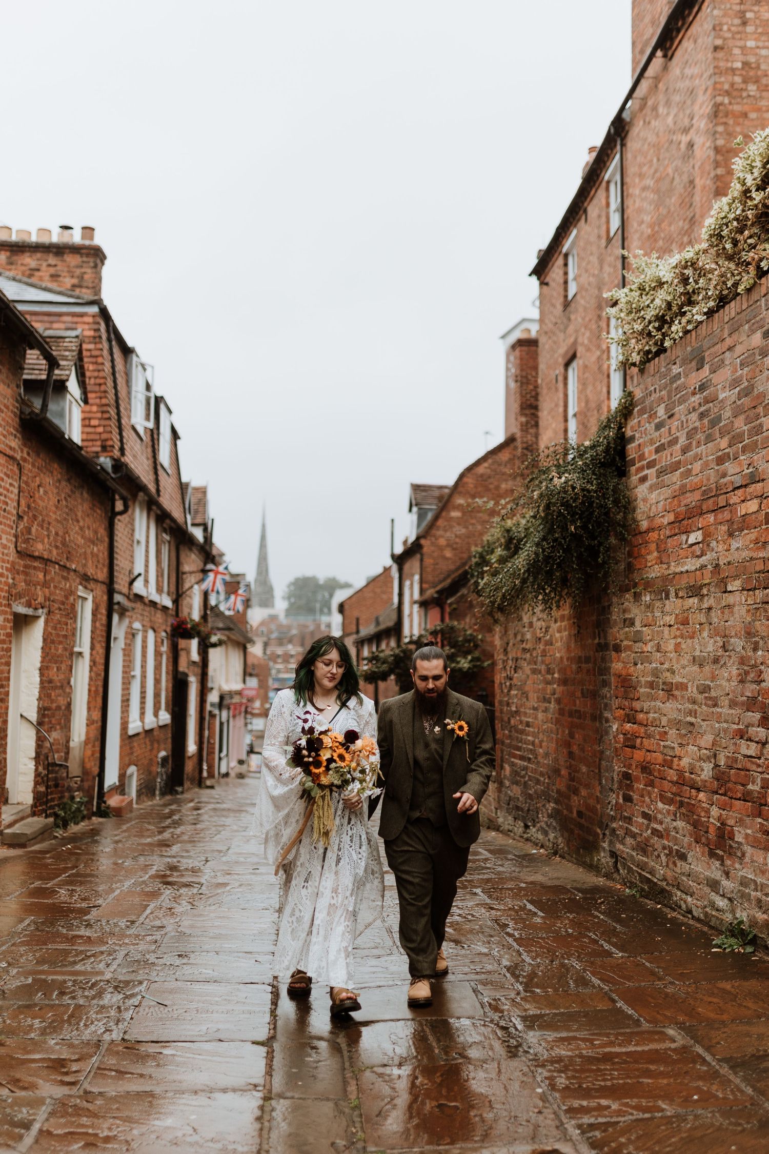 A couple walking down a rainy cobblestone street lined with historic red brick buildings in Shrewsbury.