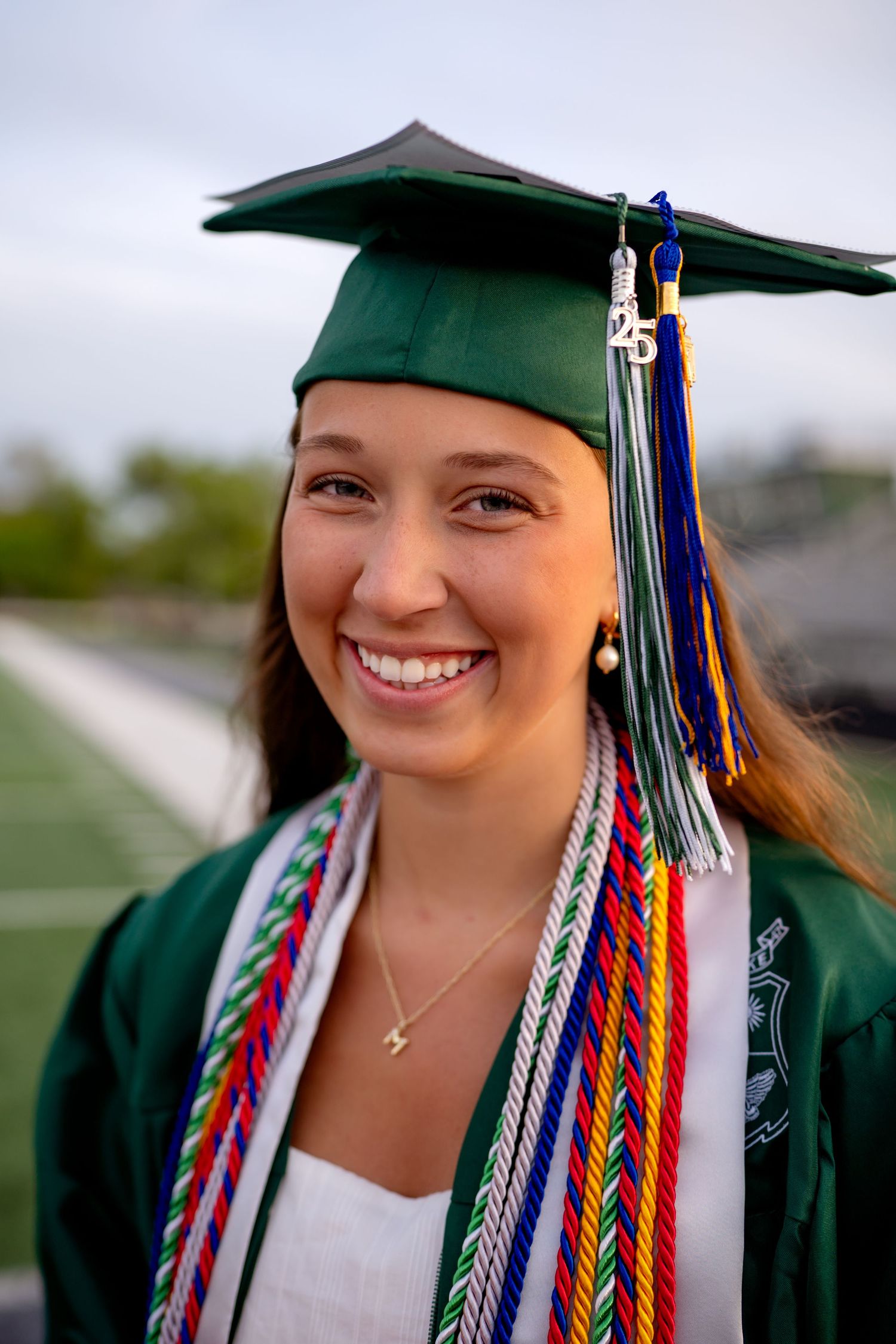 Graduate wearing a green cap and gown with colorful braided honor cords and tassels during commencement ceremony.
