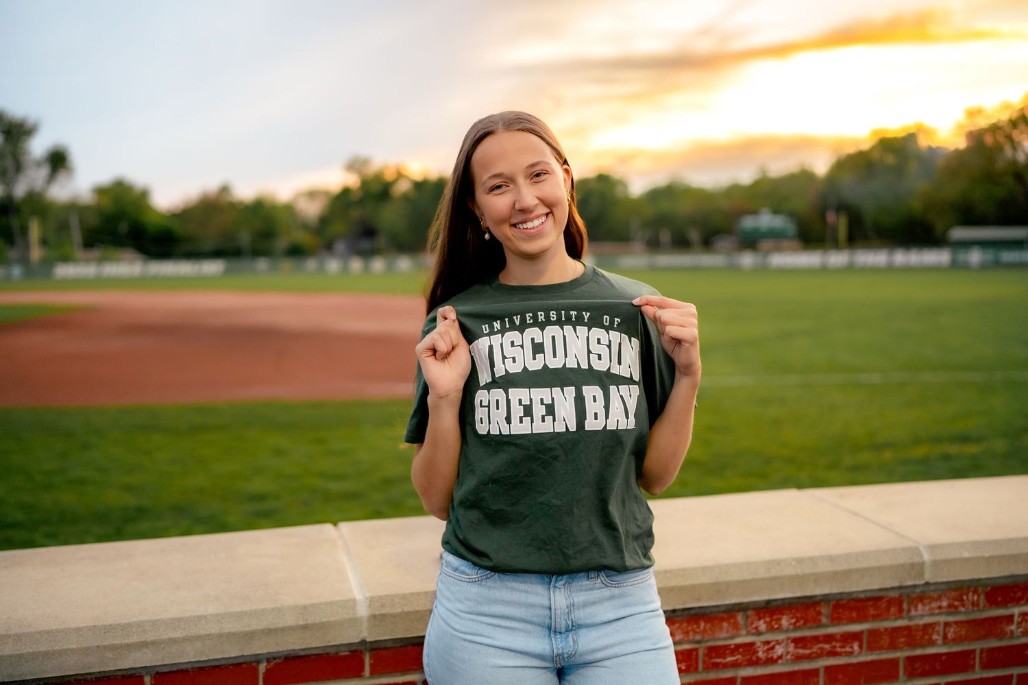A person in a dark green Wisconsin t-shirt stands smiling on a field at sunset with trees in the background.
