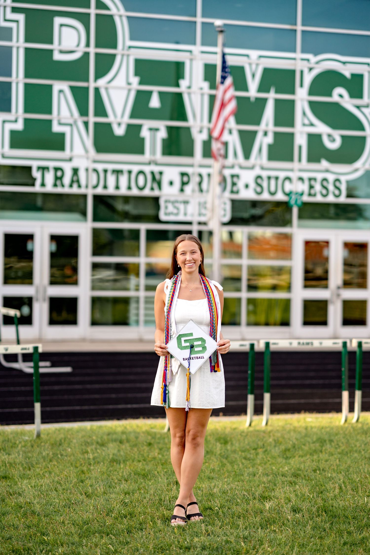 Students holding graduation certificates stand on grassy hill outside school building with green lettering.