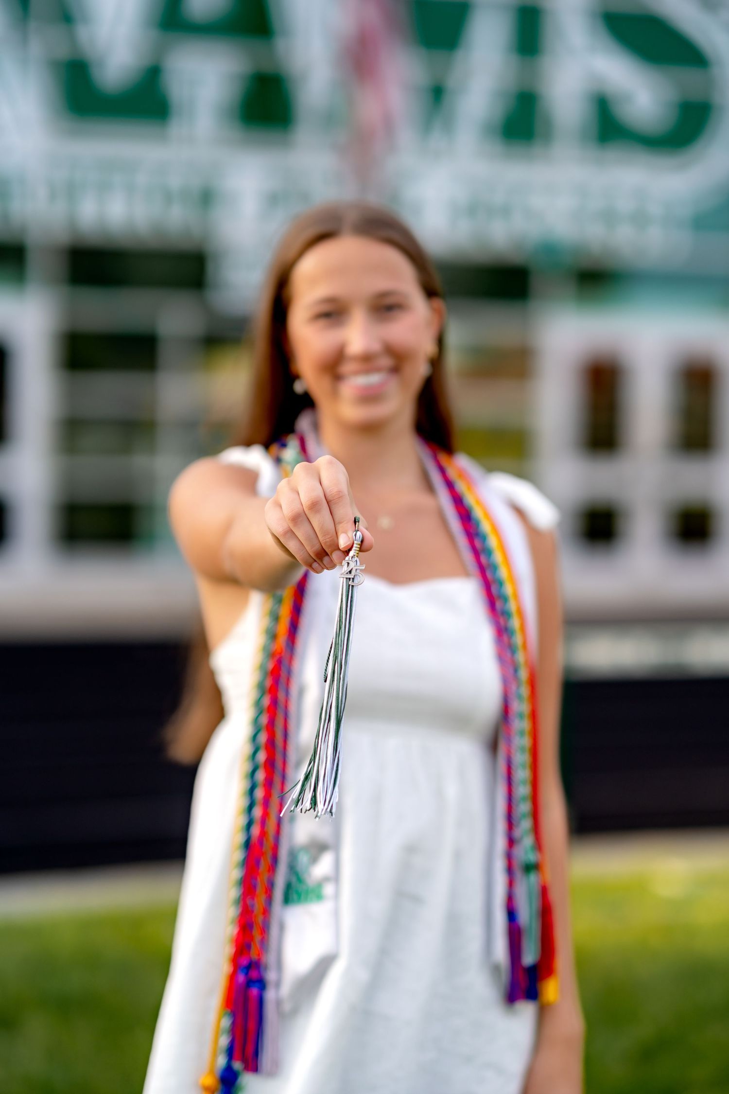 Graduate holding up their tassel in front of school building with decorative cords and white dress.