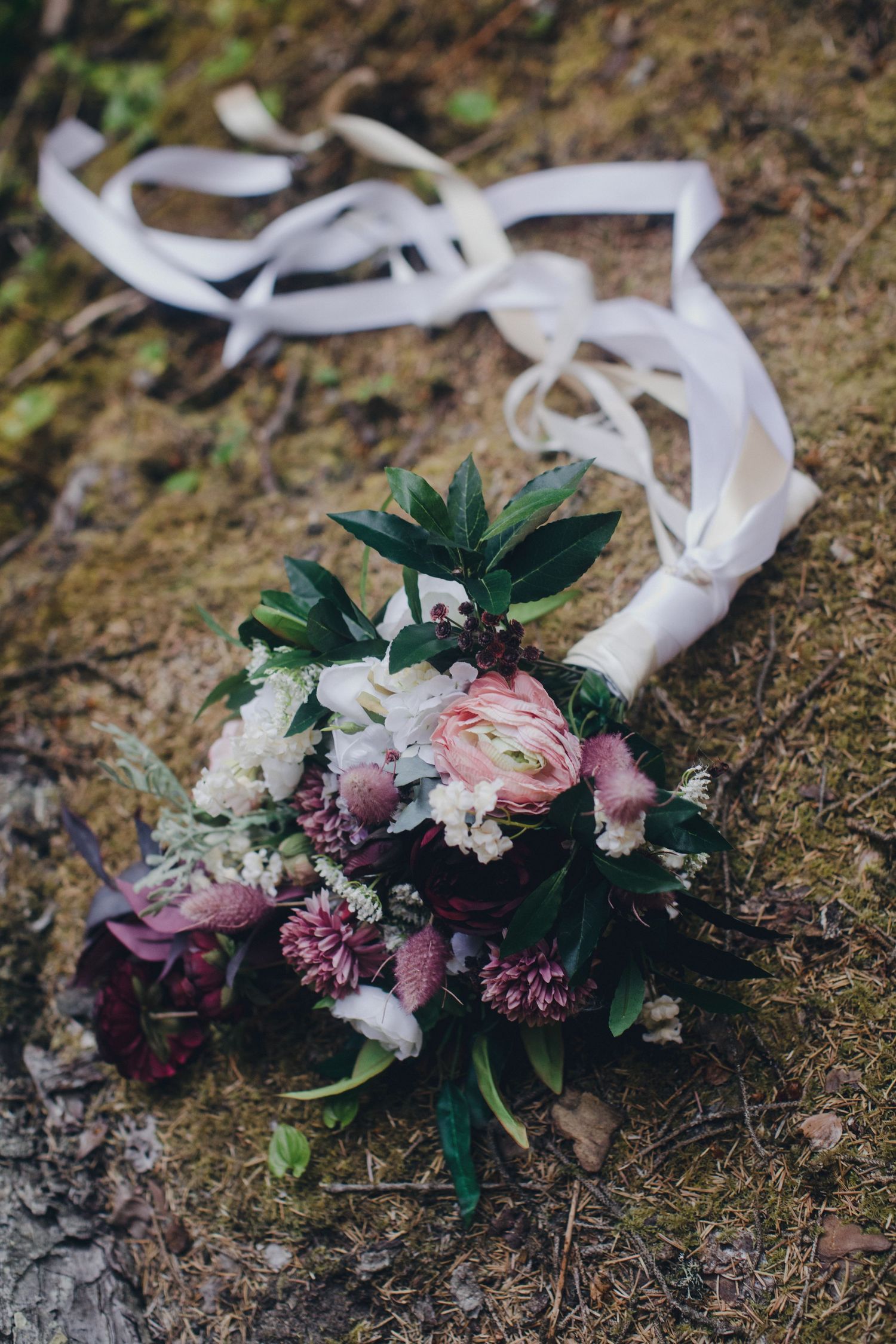 Wedding bouquet with pink and purple flowers with white ribbon on ground in moody dark lighting.