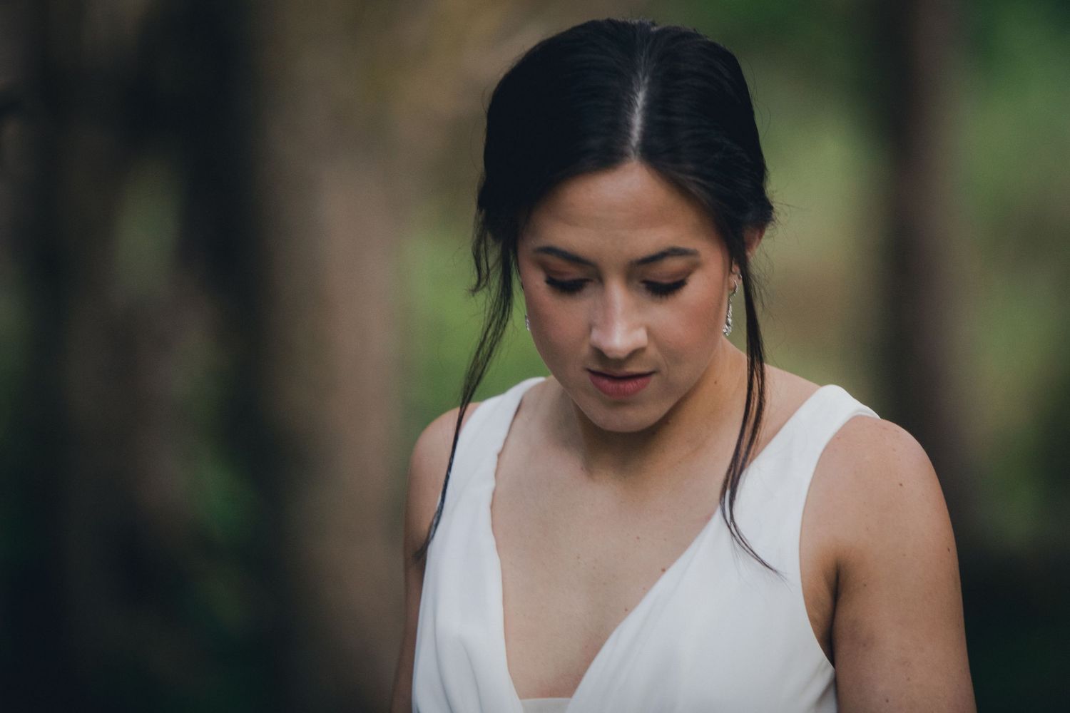 A person in a white sleeveless dress looks down thoughtfully in an outdoor setting with soft bokeh background.