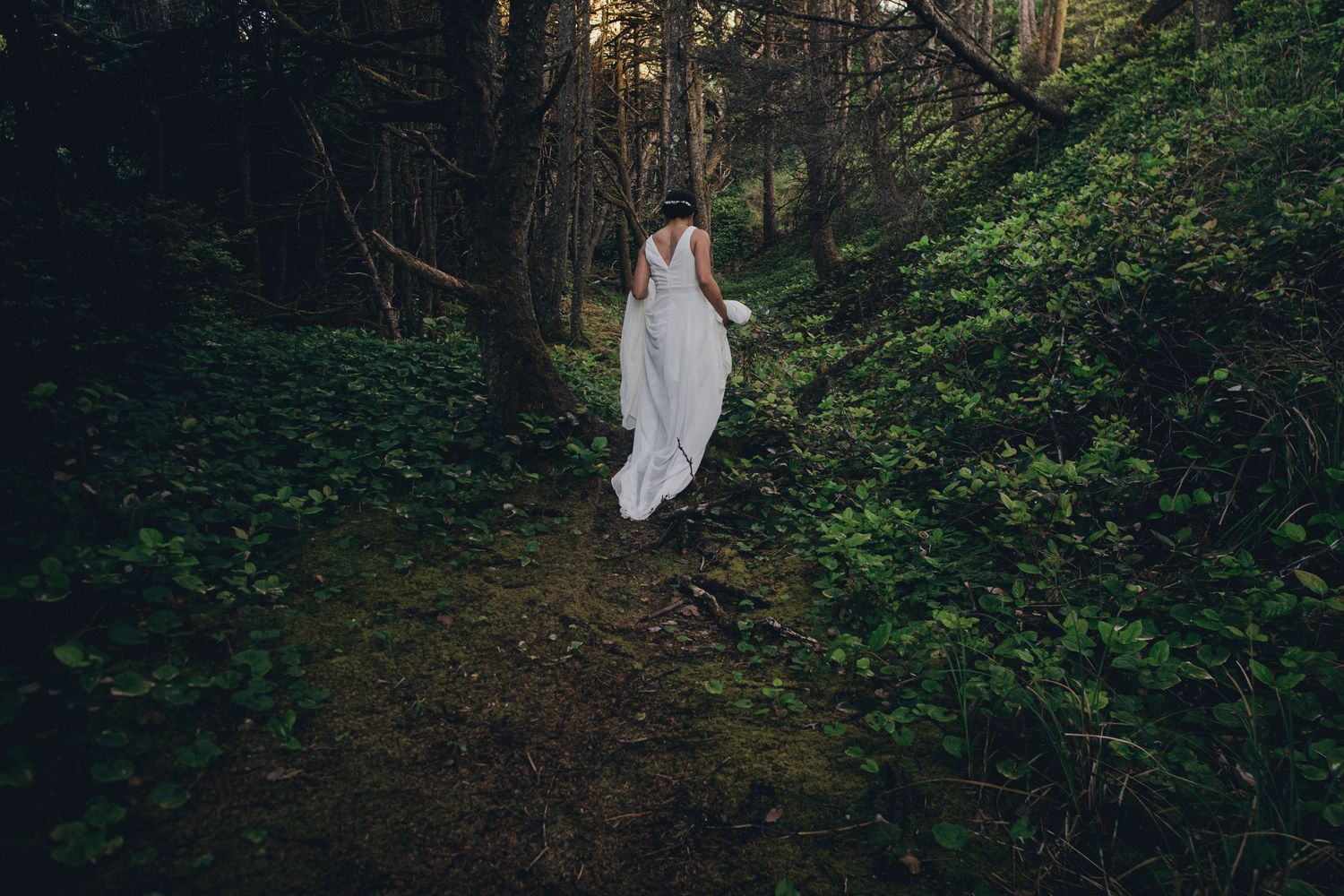 A ethereal figure in a white dress stands on a dark forest path surrounded by lush green foliage.